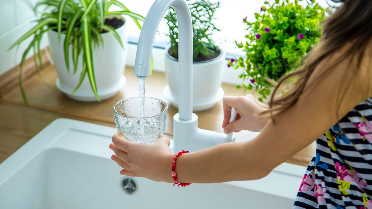 Person washing hands with water from a faucet in a kitchen setting with plants in the background.